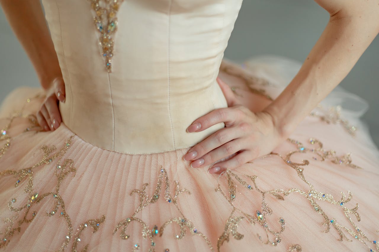 Close-up of a ballerina's detailed pink tutu with delicate embroidery, hands on hips.